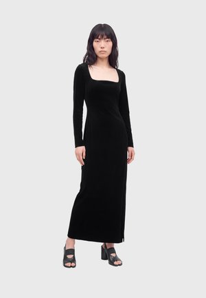 Femme aux longs cheveux foncés portant une robe maxi noire à manches longues avec encolure carrée et des sandales noires à talons ouverts, debout sur un fond blanc.