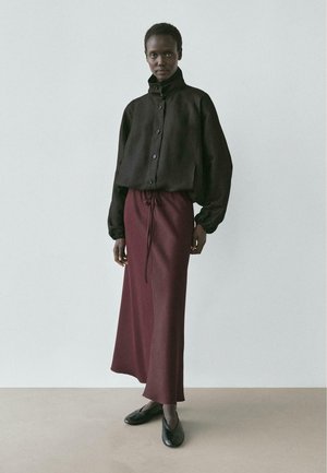 Modelka ubrana w czarną zapinaną na guziki kurtkę z wysokim kołnierzem, bordową spódnicę z troczkiem i czarne buty, stojąca na jednolitym jasnym tle.