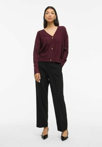 Cardigan côtelé bourgogne avec un col en V et trois boutons dorés, associé à un pantalon noir taille haute et des chaussures noires pointues.