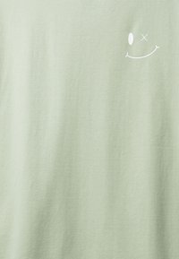 Tessuto verde pallido con un piccolo logo a faccina sorridente bianca, caratterizzato da un occhio ovale e un occhio sbarrato, nell'angolo in alto a destra.