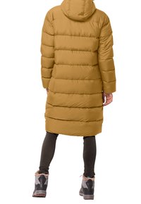 Lång pufferjacka i senapsgul med horisontella quiltade sektioner. Har en huva och kombineras med mörka leggings och grå accentstövlar.