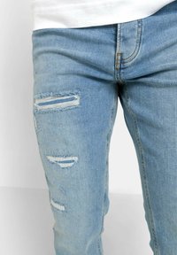 Jeans de mezclilla azul claro con múltiples áreas desgastadas, con bordes deshilachados y desgarros horizontales en los muslos, diseño de corte ajustado.