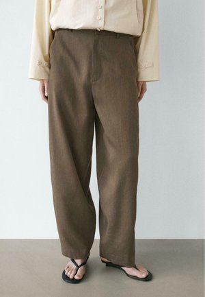 Persona con pantaloni marroni larghi, camicia beige a maniche lunghe con bottoni e sandali infradito neri in piedi su pavimento grigio.