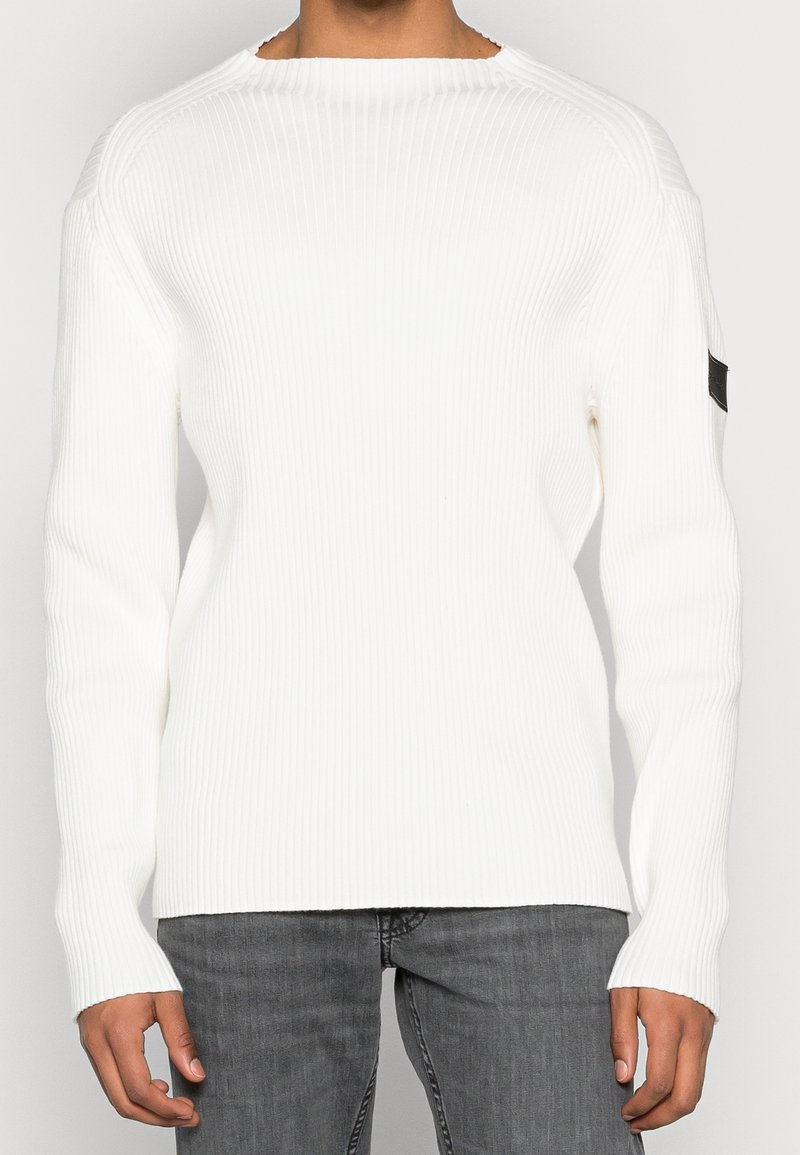 Personne portant un pull blanc côtelé à manches longues avec un patch noir sur le haut du bras gauche et un jean gris, debout sur un fond uni.