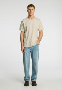 Beige cotton t-shirt with short sleeves, paired with light blue denim jeans. Model stands upright, wearing brown shoes. Simple, casual design.