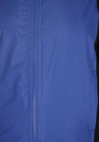 Giacca leggera blu con texture liscia, chiusura frontale con zip e accenti neri a contrasto sui lati.
