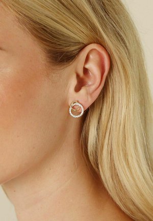 Guldsmycken i form av hoops med sammanlänkade cirkulära mönster, en prydd med små klara kristaller, som visar en polerad metallisk finish.