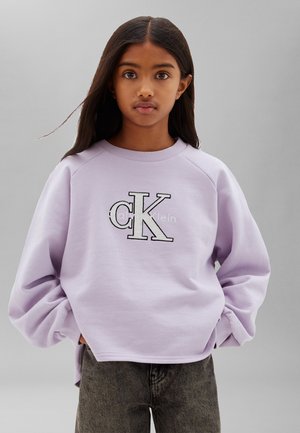 Junges Mädchen mit langen dunklen Haaren trägt einen lavendelfarbenen Calvin Klein Pullover und graue Jeans, steht vor einem schlichten hellgrauen Hintergrund.