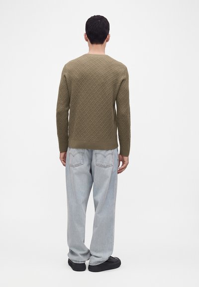 Homme debout, dos à la caméra, portant un pull olive texturé, un jean ample bleu clair et des chaussures noires sur un fond blanc uni.