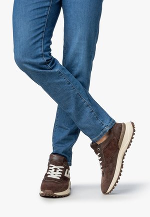 Bruine suède sneakers met witte veters en rubberen zolen, gecombineerd met blauwe denim jeans. De sneakers hebben een gestructureerd ontwerp en subtiele branding.
