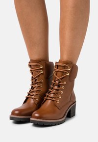 Bottes chelsea en cuir marron avec un bout arrondi, des œillets dorés et des lacets. Présentent un talon épais et un col rembourré pour le confort.
