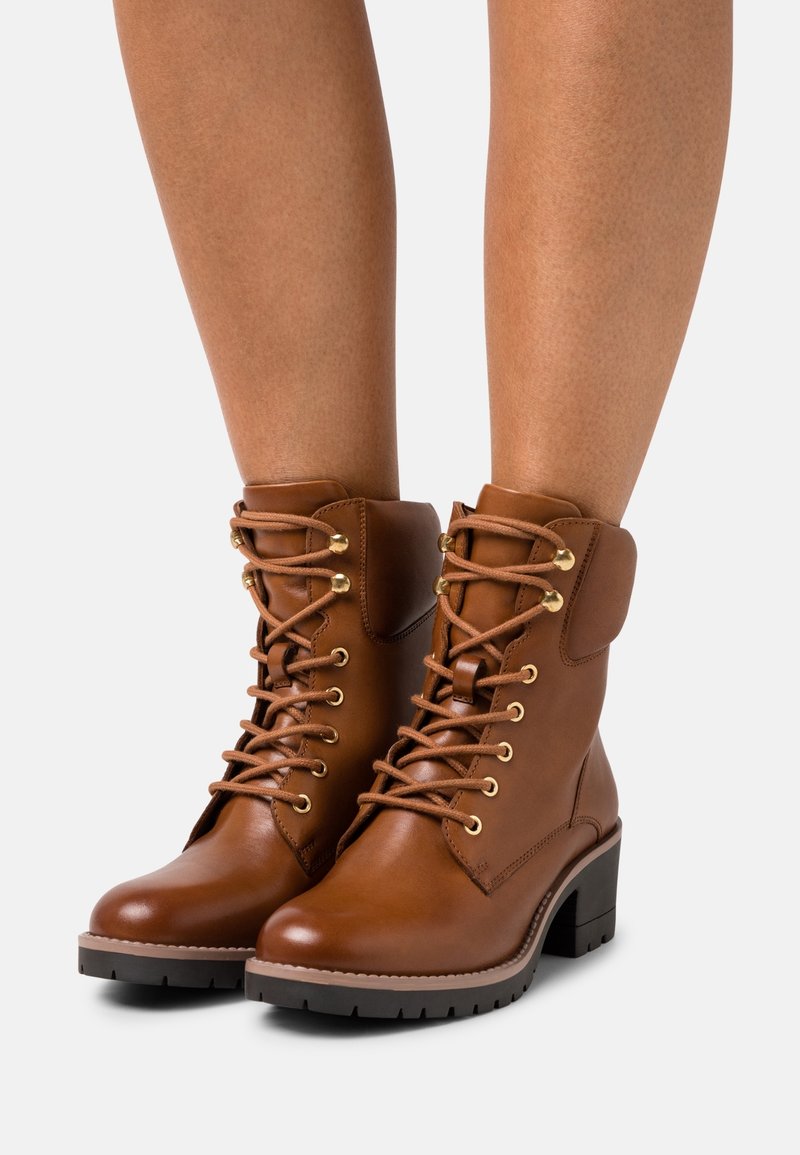Bottes chelsea en cuir marron avec un bout arrondi, des œillets dorés et des lacets. Présentent un talon épais et un col rembourré pour le confort.