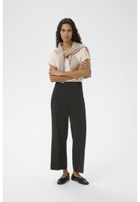 Svarta byxor med vida ben, ljusbeige skjorta och en beige stickad tröja knuten runt axlarna. Enkel design; lättviktsmaterial.