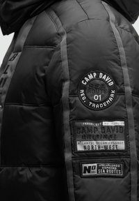 Schwarze Pufferjacke mit gestepptem Muster, grauen Streifen und mehreren Stoffflicken mit Text, darunter "Camp David" und Richtungsbeschriftungen.