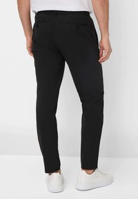 Zwarte broek met een slim fit, twee achterzakken met knoopsluitingen, gecombineerd met witte sneakers. Glad materiaal, strakke lijnen.
