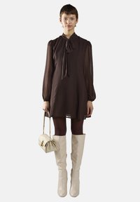 Braunes Chiffonkleid mit einem Bindekragen, langen Ärmeln und einer fließenden Silhouette. Kombiniert mit kniehohen, gläsernen weißen Stiefeln und einer kleinen Handtasche.