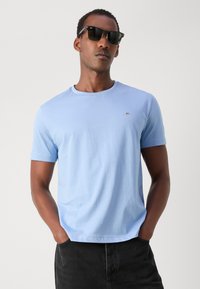 Light blue cotton t-shirt with a round neckline, short sleeves, and a small embroidered logo on the chest, paired with black denim pants.