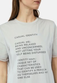 Jasnoszara koszulka z krótkim rękawem i okrągłym dekoltem, z nadrukowanymi w czerni definicjami słów "casual" i "identity".