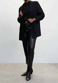 Blazer oversize nero su un dolcevita nero, abbinato a pantaloni in pelle nera a vita alta e stivaletti neri con punta a contrasto e dettagli metallici.