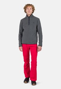Pullover in pile grigio con colletto alto e zip a quarti, abbinato a pantaloni rossi vivaci. Scarpe nere completano l'outfit.