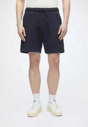 Mężczyzna ubrany w czarne szorty ze sznurkiem, białe skarpetki i białe sneakersy Diesel, stojący na jednolitym białym tle.