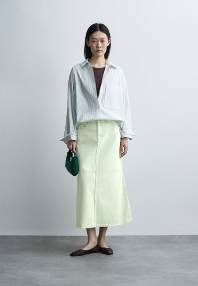 Femme debout portant une chemise blanche surdimensionnée, un haut marron, une jupe midi vert clair, des ballerines foncées, tenant un petit sac à main vert sur un fond uni.