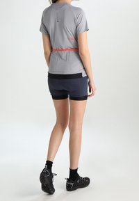 Camisa atlética gris de manga corta con un acento rojo y pantalones cortos oscuros ajustados. Modelo usando zapatos de escalada negros, de pie sobre una superficie lisa.