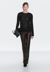 Kvinde iført sort langærmet bluse, mørkebrune læderbukser, sorte støvler og bærende en sort håndtaske mod en hvid baggrund.