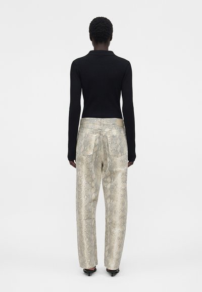 Άτομο που φοράει μαύρο τοπ με μακριά μανίκια και παντελόνι με φαρδιά, ψηλή μέση και γκρι σχέδιο φίδι, στέκεται με την πλάτη προς τα εμπρός σε απλό φόντο.