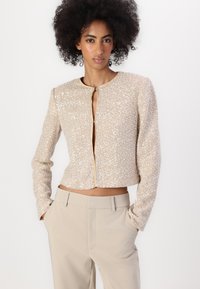 Giacca corta con perline in beige, maniche lunghe, scollatura rotonda e chiusura con zip frontale; abbinata a pantaloni chiari.