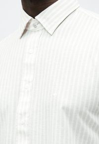 Weiß gestreiftes Hemd mit Button-Down-Kragen. Der Stoff hat eine glatte Textur. Mit kleinem Logo auf der linken Seite und dezenten Knöpfen.