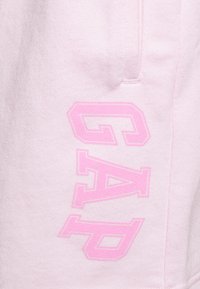 Pantaloni della tuta rosa chiaro con una texture morbida, caratterizzati da un grande logo "GAP" rosa in rilievo sulla gamba sinistra e da una tasca laterale.