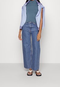 Una modella indossa un dolcevita aderente color teal, jeans a palazzo larghi blu denim e sandali bianchi con suole nere. Una camicia a righe blu è appesa.