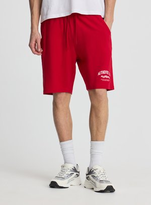 Uomo che indossa pantaloncini rossi con testo bianco sulla coscia sinistra, calzini bianchi, scarpe da ginnastica bianche e una maglietta bianca, in piedi con le mani nelle tasche.