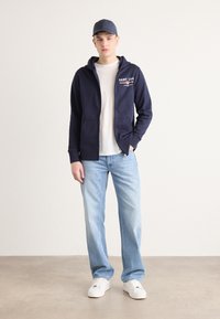Sweat à manches zippées marine avec poches avant, texte graphique blanc et un T-shirt clair en dessous. Associé à un jean bleu clair et des baskets blanches.