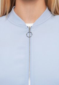 Blouson aviateur bleu clair avec col côtelé et une fermeture éclair argentée munie d'un tirette circulaire. Texture de tissu lisse, design simple.