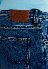 Blue denim jeans with a brown leather patch on the back, featuring stitching details, a classic five-pocket design, and a smooth texture.