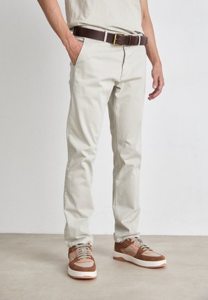 Homme portant un pantalon chino beige clair, ceinture en cuir marron, et baskets bicolores beige et marron, debout avec une main dans la poche.
