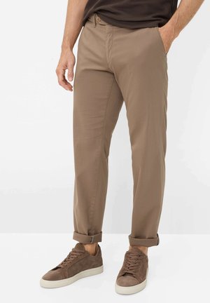 Homme portant un pantalon marron clair retroussé avec des baskets en daim marron assorties sur fond blanc.