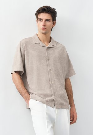 Νεαρός άνδρας που φοράει μπεζ πουκάμισο με κοντά μανίκια και λευκό παντελόνι, στέκεται με το ένα χέρι στην τσέπη, κοιτάζοντας ελαφρώς προς τα αριστερά.