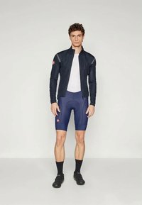 Marine sykkeldress med glidelås, lange ermer; hvit mesh-baselayer; marineblå polstrede shorts; svarte sokker; sykkelsko med spenner.