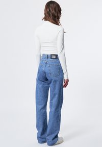 Leichtes weißes Langarmoberteil kombiniert mit hochgeschnittenen blauen Jeans aus Denim, die einen geraden Schnitt und ein markiertes Rückenteil haben.