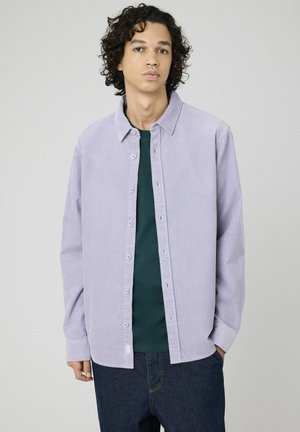 Chemise en corduroy violet clair avec fermeture à boutons et col, portée avec un t-shirt vert en dessous, associée à un jean en denim noir.