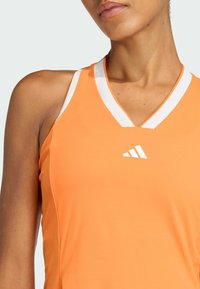 Débardeur de sport orange avec un col en V, comportant des accents en maille blanche et un logo blanc sur la poitrine, fabriqué en tissu lisse et extensible.