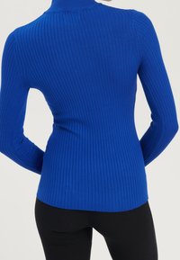 Rippstrick-Turtleneck mit langen Ärmeln in leuchtendem Blau, eng anliegendes Design mit strukturiertem Stoff. Kombiniert mit schwarzen eng anliegenden Hosen, die am Saum sichtbar sind.