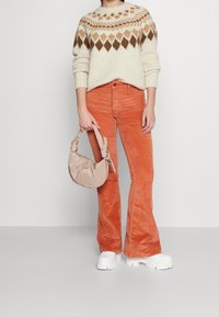 Pull en maille avec un motif losange beige et marron, associé à un pantalon évasé en velours côtelé orange et un sac beige doux. Chaussures blanches épaisses.