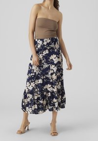 Kvinna med en ärmlös taupe topp och en högmidjad marinblå midikjol med vita blommönster samt nudefärgade klacksandaler.