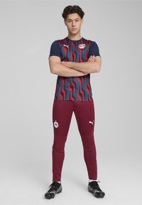 Vörös és tengerészkék sportegyenruha, hullámzó mintával, rövid ujjú póló logóval, bordó szűk nadrággal, oldalán díszítéssel. Fekete sportcipő viselve.