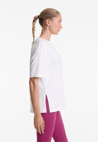 Nő fonott lófarokkal, fehér, rövid ujjú oldalt hasított felsőt és bíbor leggingset viselve, jobbra néz, egyszínű háttér előtt.