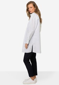 Camicia button-up bianca in cotone con colletto alto, fessure laterali e dettagli gialli sul retro, abbinata a jeans scuri e sneaker bianche.